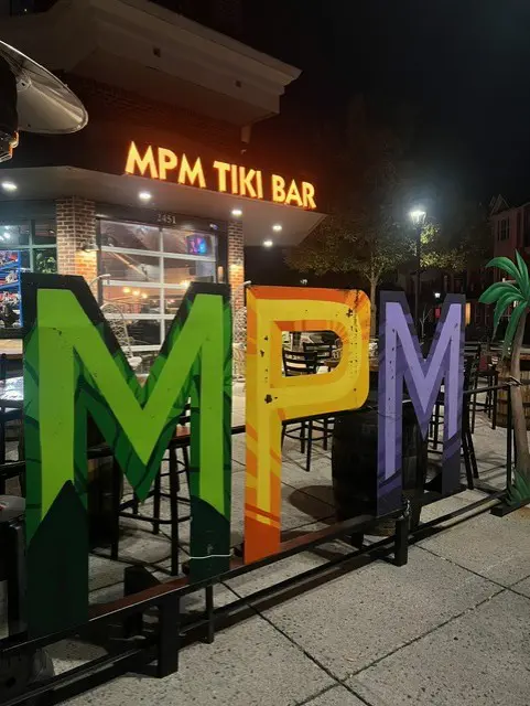 Large colorful freestanding letters spelling MPM in green, orange and purple on an outdoor patio with tiki palm decor and bar stools, illuminated at night in front of the glowing MPM TIKI BAR sign on the Short Pump restaurant&amp;#39;s façade