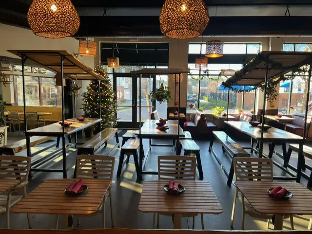 Interior of MPM Tiki Bar in Short Pump with tiki-style woven pendant lights, wooden tables and benches set with napkins, booth seating by large windows, holiday tree and greenery, and sunlight streaming into the cozy Thai-inspired dining and cocktail space