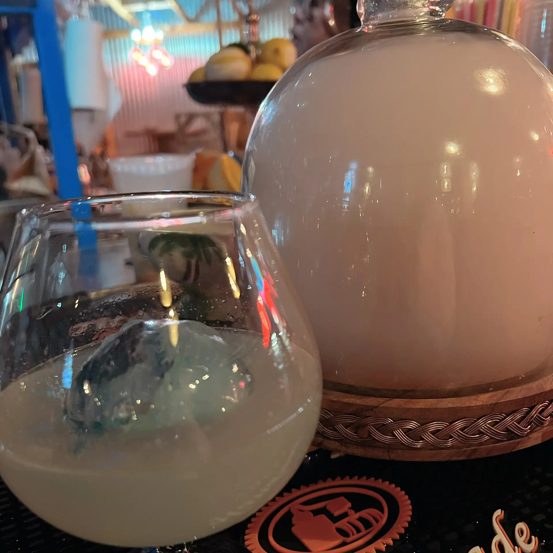 A close-up of a tiki-style cocktail in a round glass with a single large ice sphere beside a smoke-filled glass cloche on a bar mat, colorful Short Pump tiki bar lights and fruit display blurred in the background